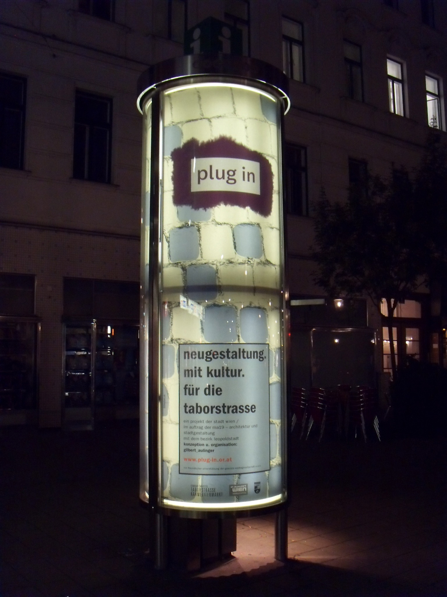 Night. One side of large glass advertising pillar in the city. Poster illuminated inside. Illustrated cobblestone floor allover. Upper half logo and lower half textbox overlayed.