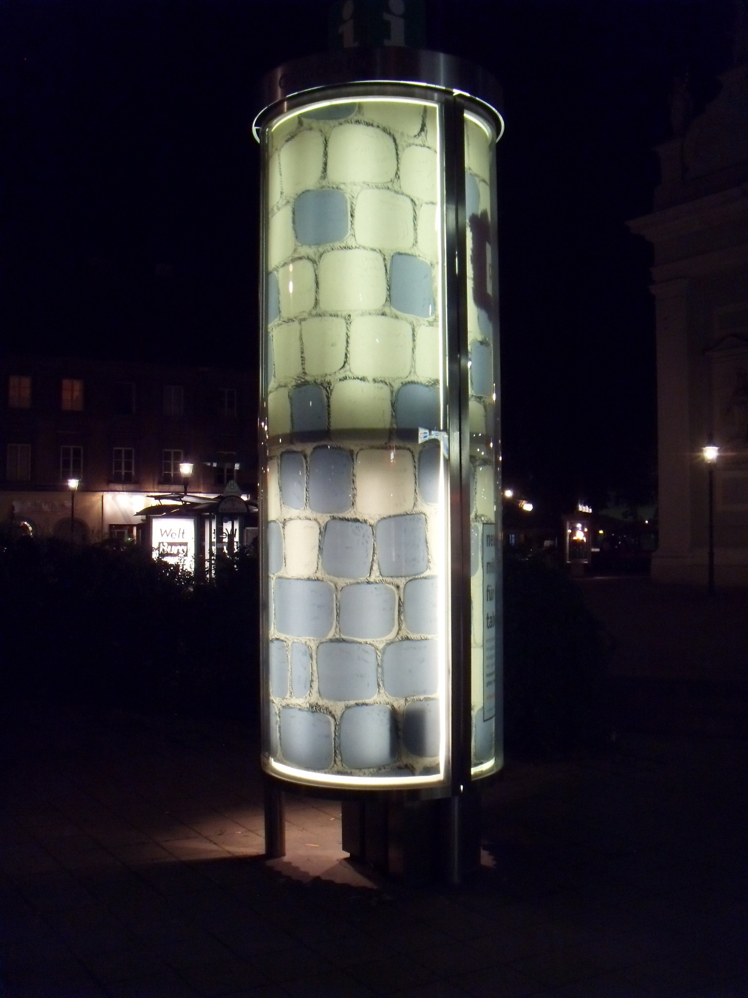 Other side of large advertising pillar. Poster illuminated inside. Illustrated cobblestone floor allover.