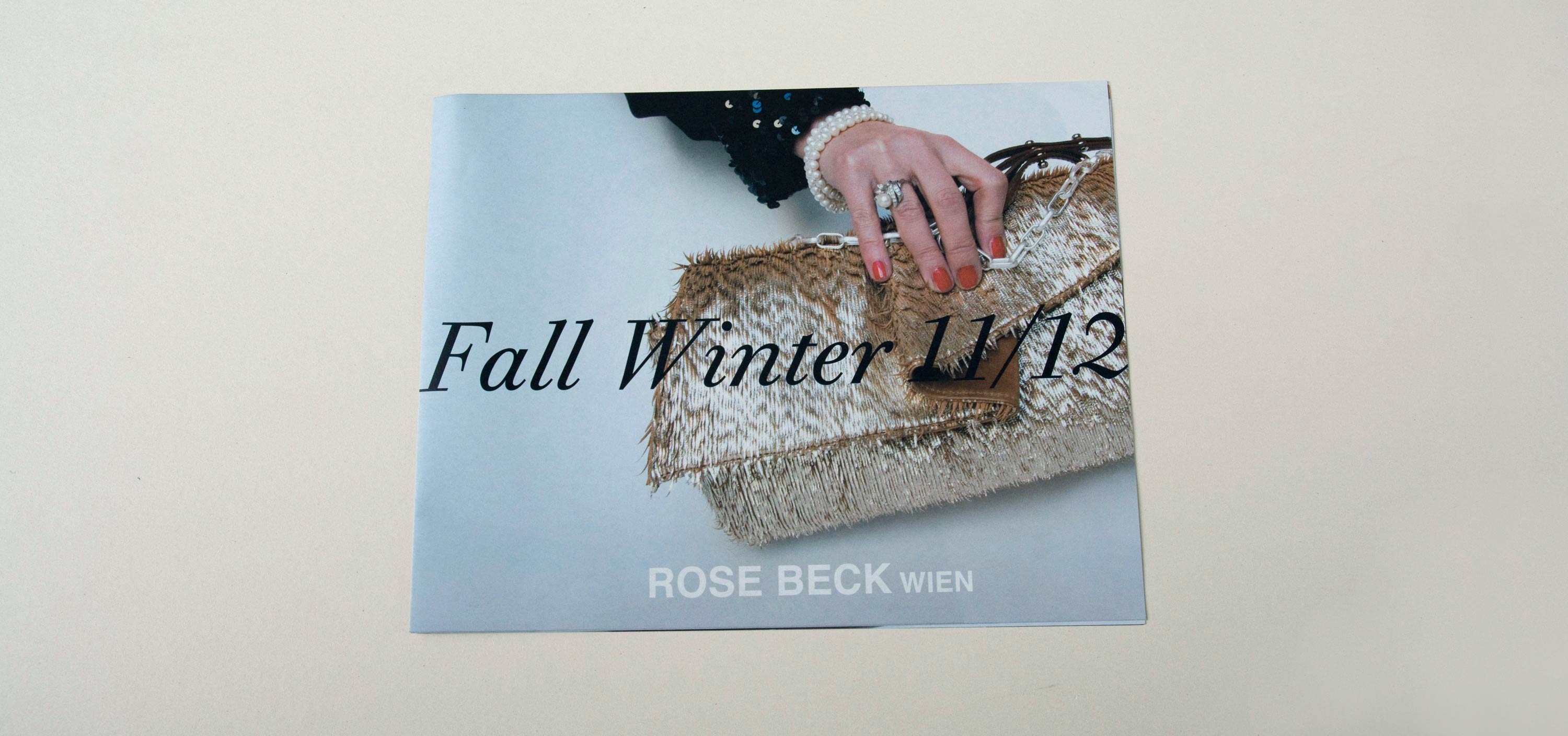Cover catalogue. Full-page close-up photo of hand holding handbag. Large title in script font overlayed. Logo at bottom overlayed.