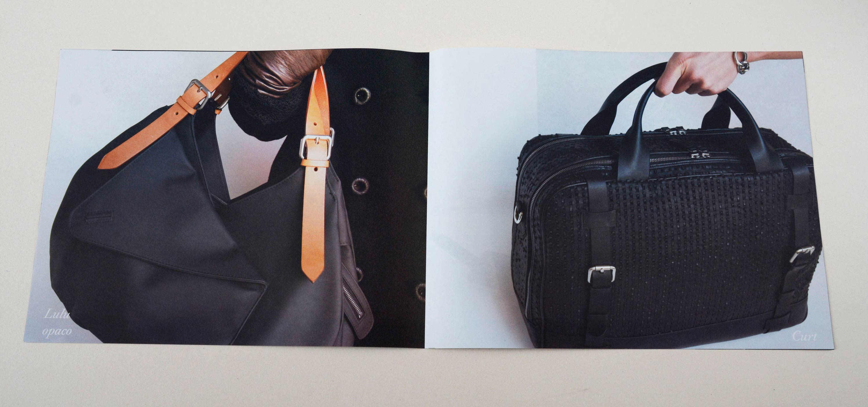 Double page with full-page photos. Script font overlayed at bottom. Left: Close-up of hand holding handbag. Right: Close-up of bag standing on neutral floor. Hand holds the handles.
