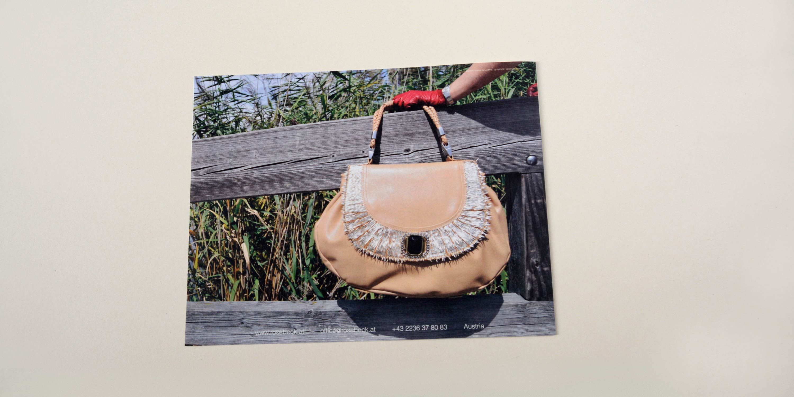 Back catalogue. Full-page photo hand holding large handbag in front of wooden railing. Reeds in the background. Row of small text at bottom overlayed.