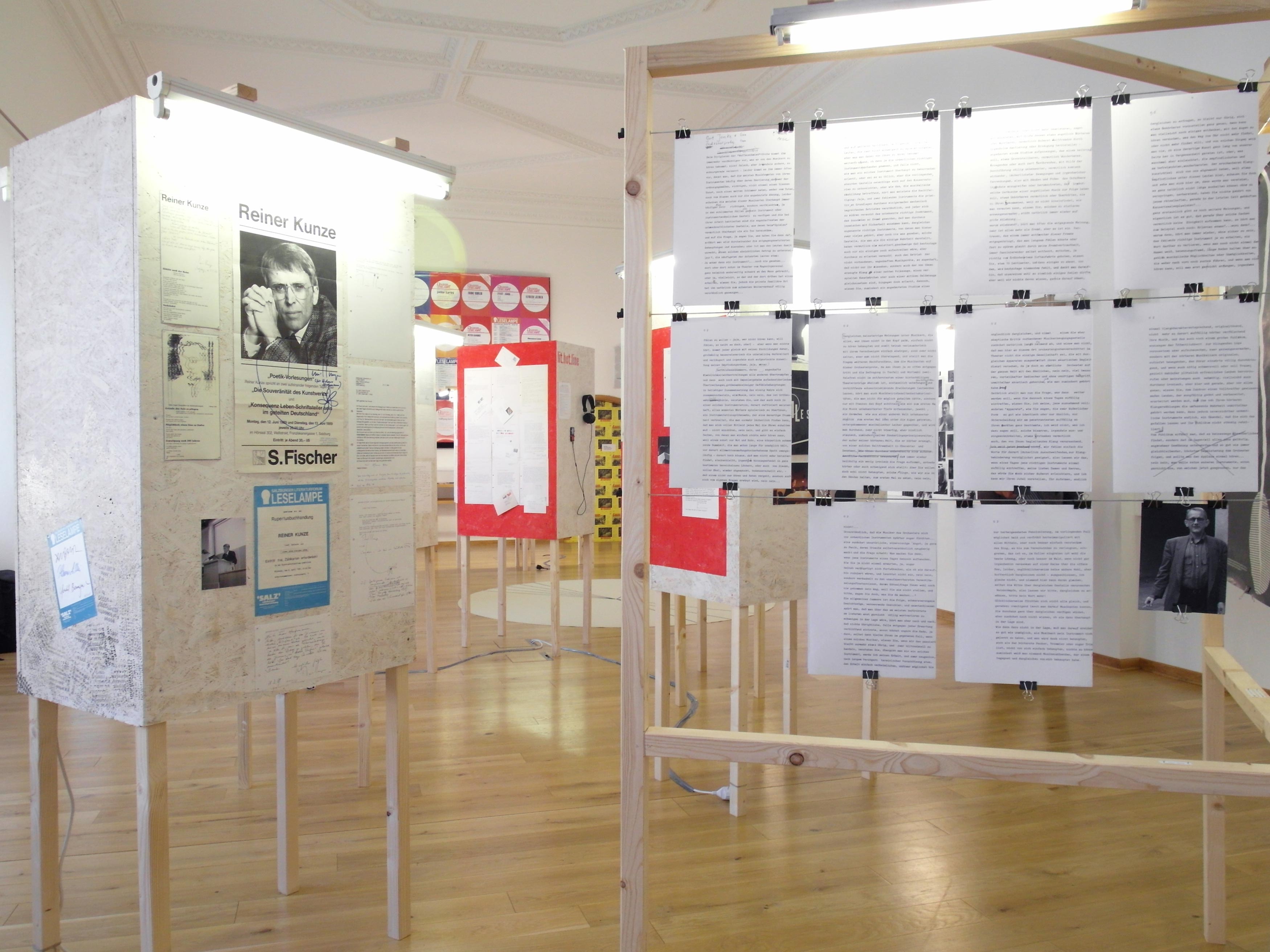 Room with several wooden cuboids mounted on 4 squared timber. Sheets of paper with texts and images are mounted on the cuboids. Inside a wooden frame metal cords are stretched. Sheets of paper with text are clipped to the cord.