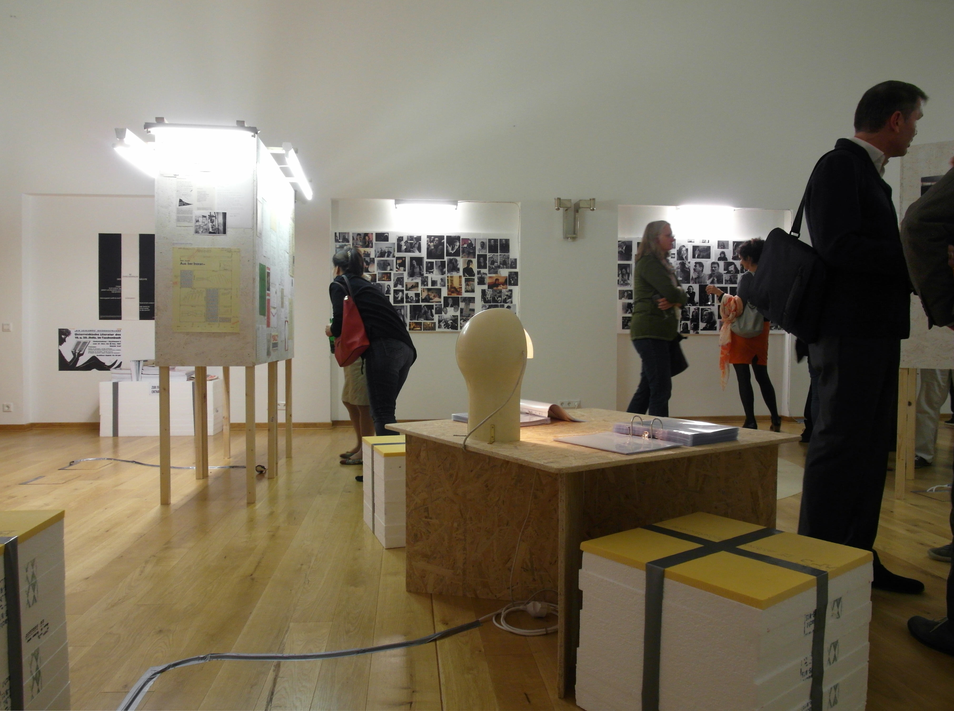 People in a room. Looking at wall of photos and sheets of paper fixed on a cuboid.