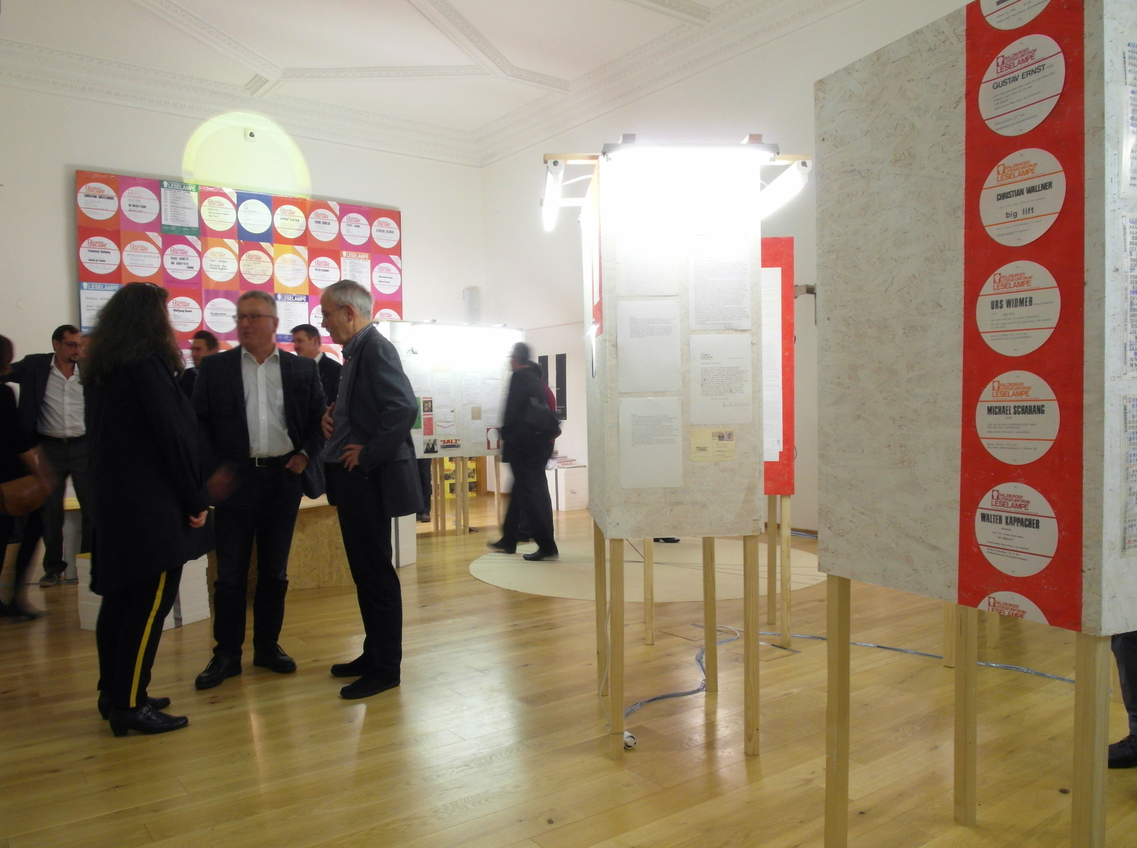 Row of cuboids in the room. Several graphical images and sheets with text are hung up at them. Beside people talking to each other.