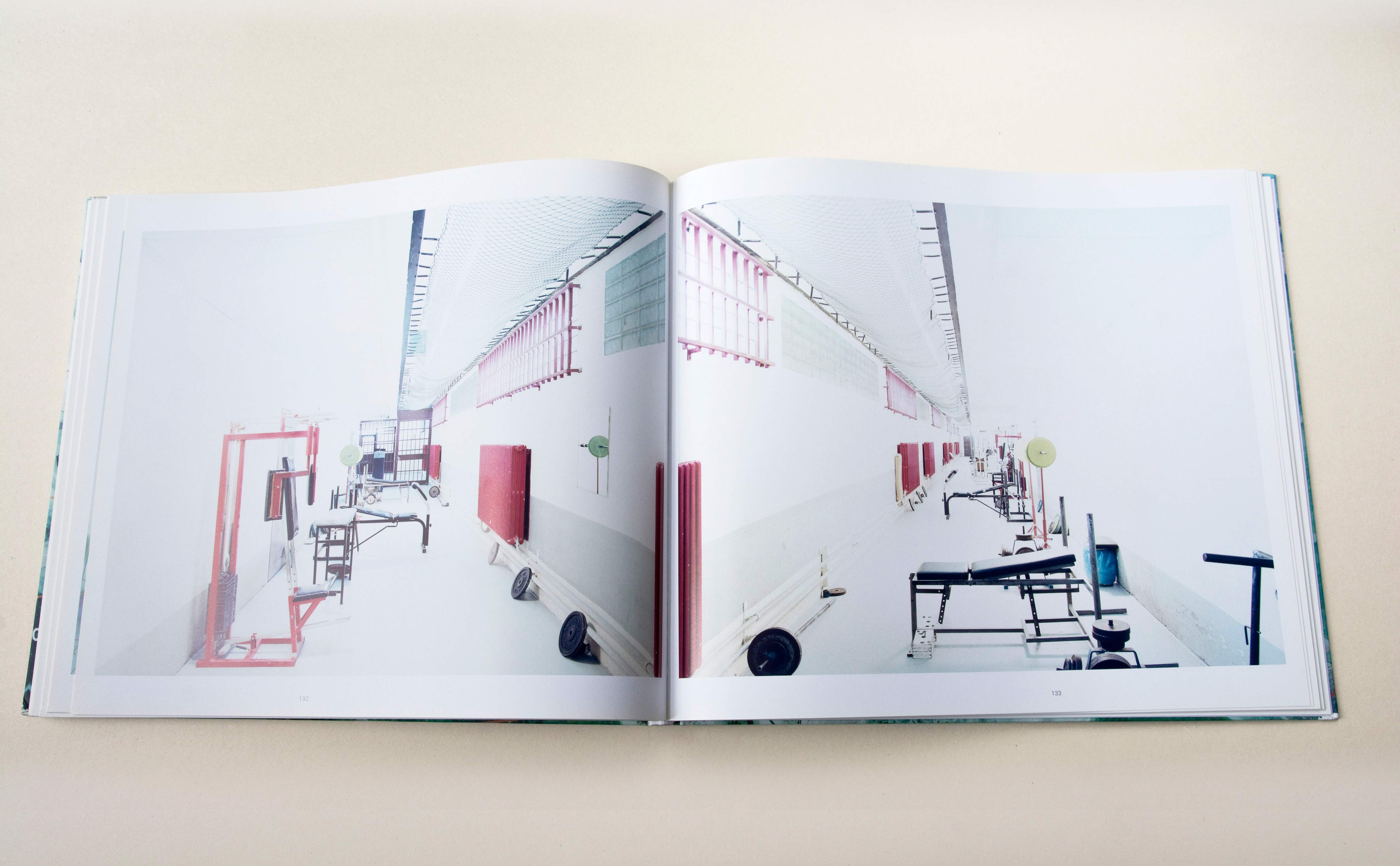 Double page. Large photo with white space around on each page. Left: Corridor in a prison. Fitness devices standing at the side. Right: Similar shot of corridor mirrorerd.