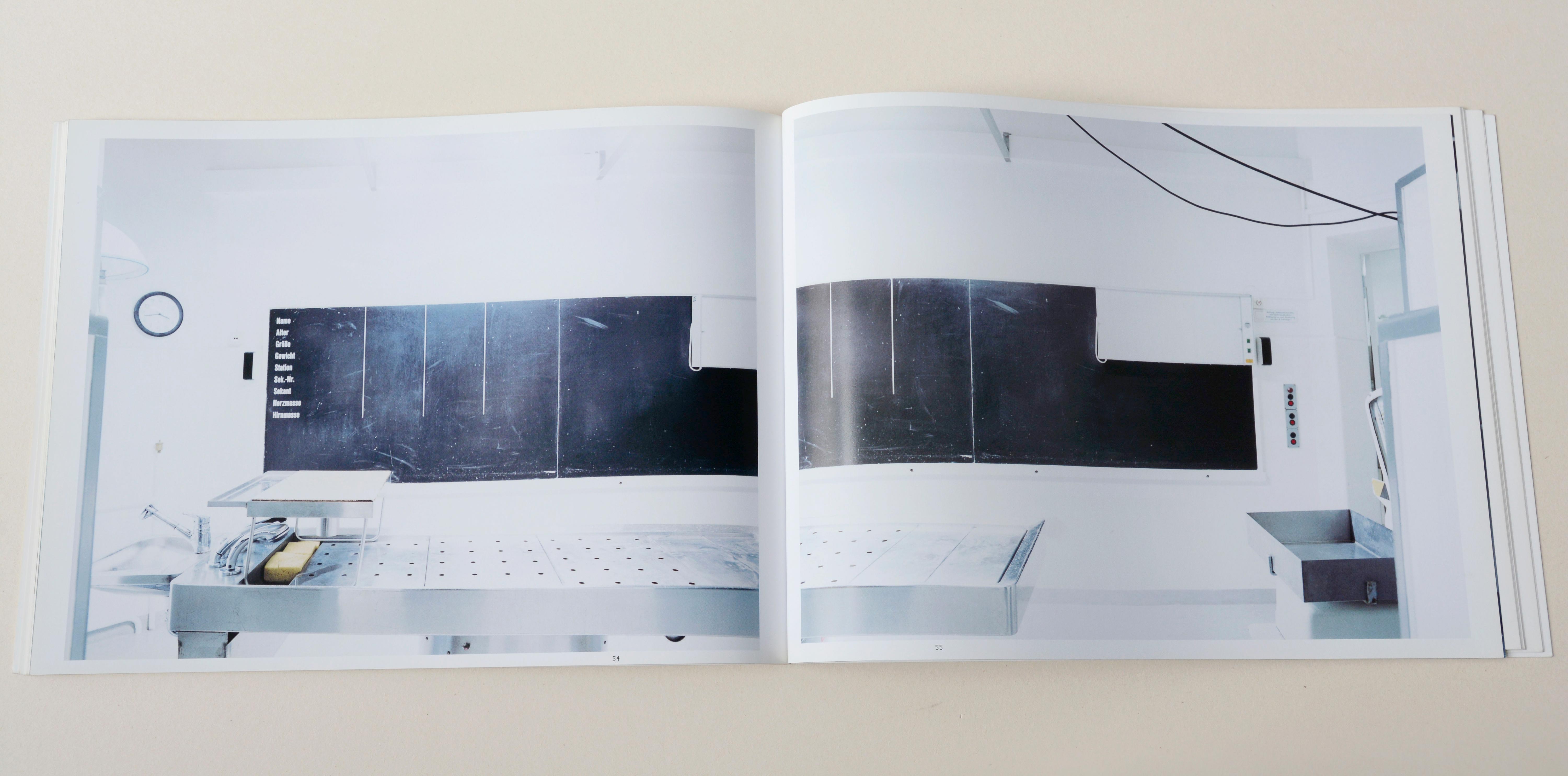 Double page. Large photo with white space around on each page. Left: White sterile room. Metal stretcher in front. Blackboard on wall in back. Right: Partly same and follow-up section of left photo.