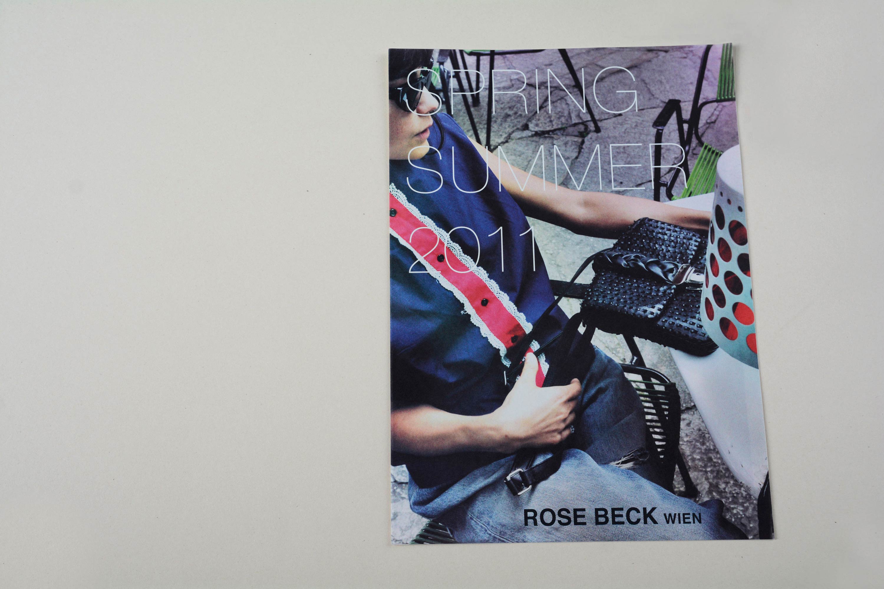 Lookbook cover front. Full-page close-up of woman sitting at a table outside in a bar. Large title in thin grotesque font overlayed. Logo overlayed at bottom.