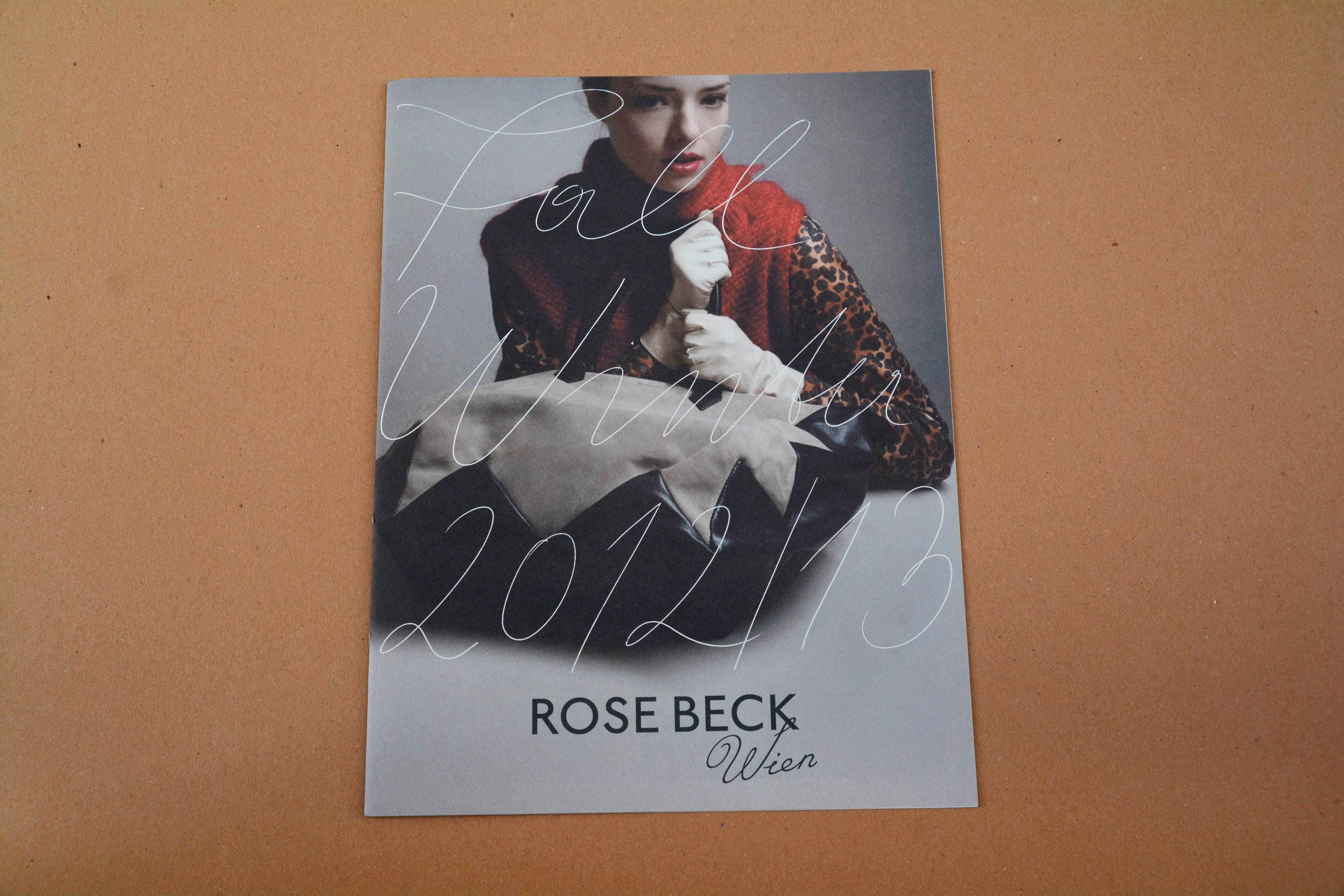 Cover catalogue. Full-page photo of handbang lying on white table. Woman behind holds the handles. Title overlayed stretches over whole cover in thin script font. Logo at bottom.