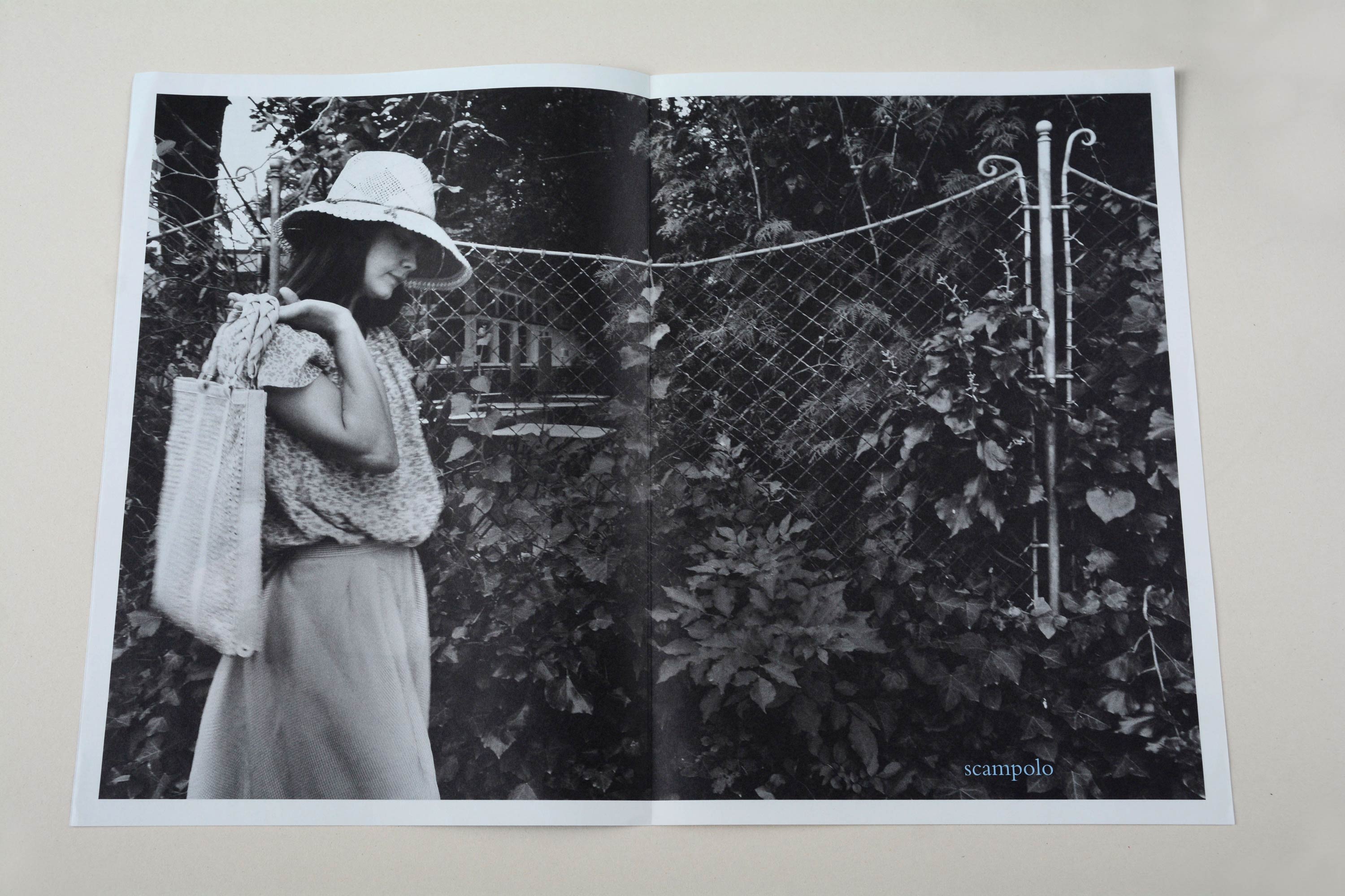 Double page. Full-page b/w photo with white space around stretches over both pages. Woman walking in front of iron grid fence. Bushes behind fence. Small font at bottom overlayed.