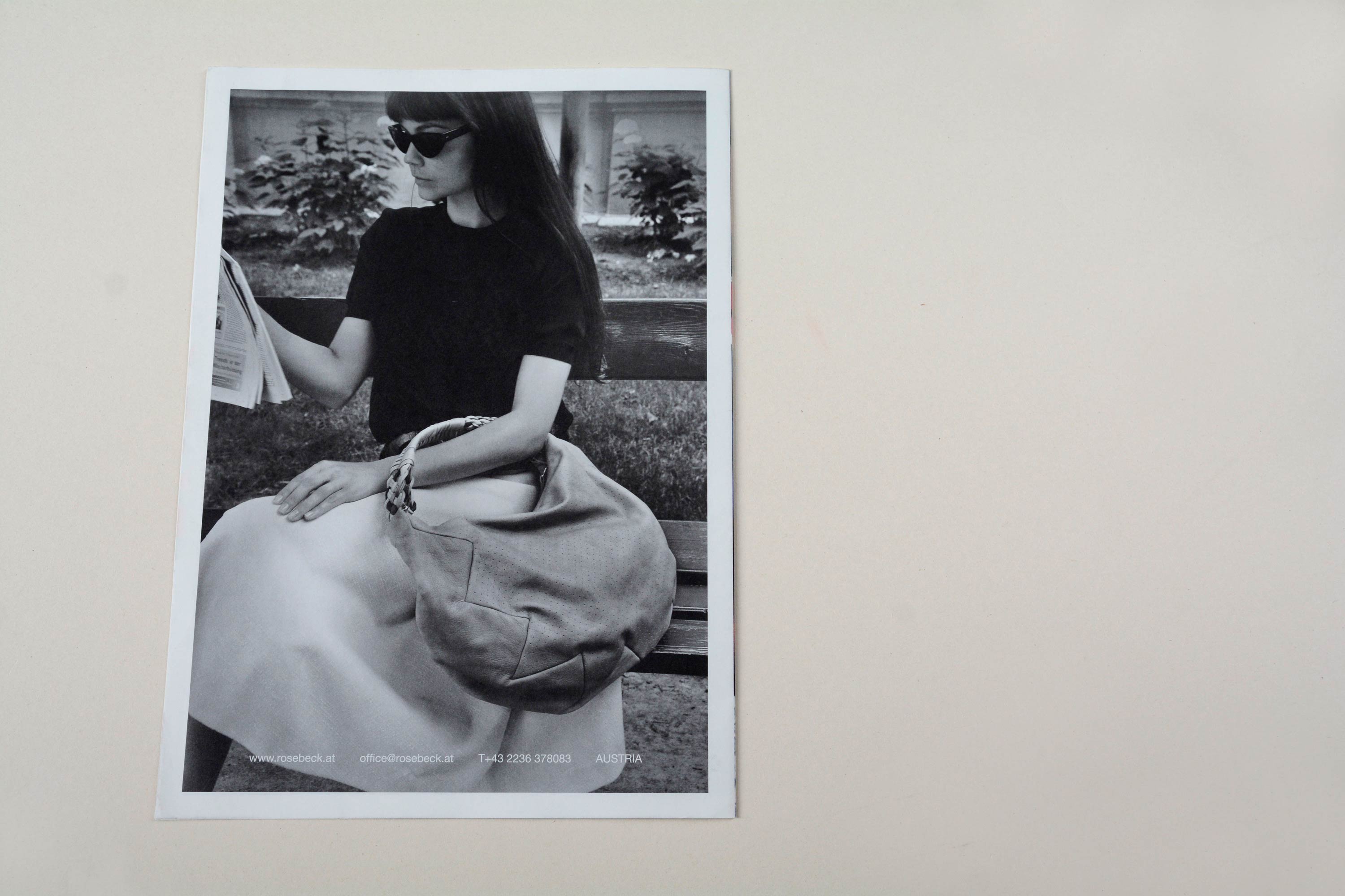 Back catalogue. Full-page b/w photo with white space around. Woman sitting on park bench with large handbag around her arm. Row of small text at bottom overlayed.
