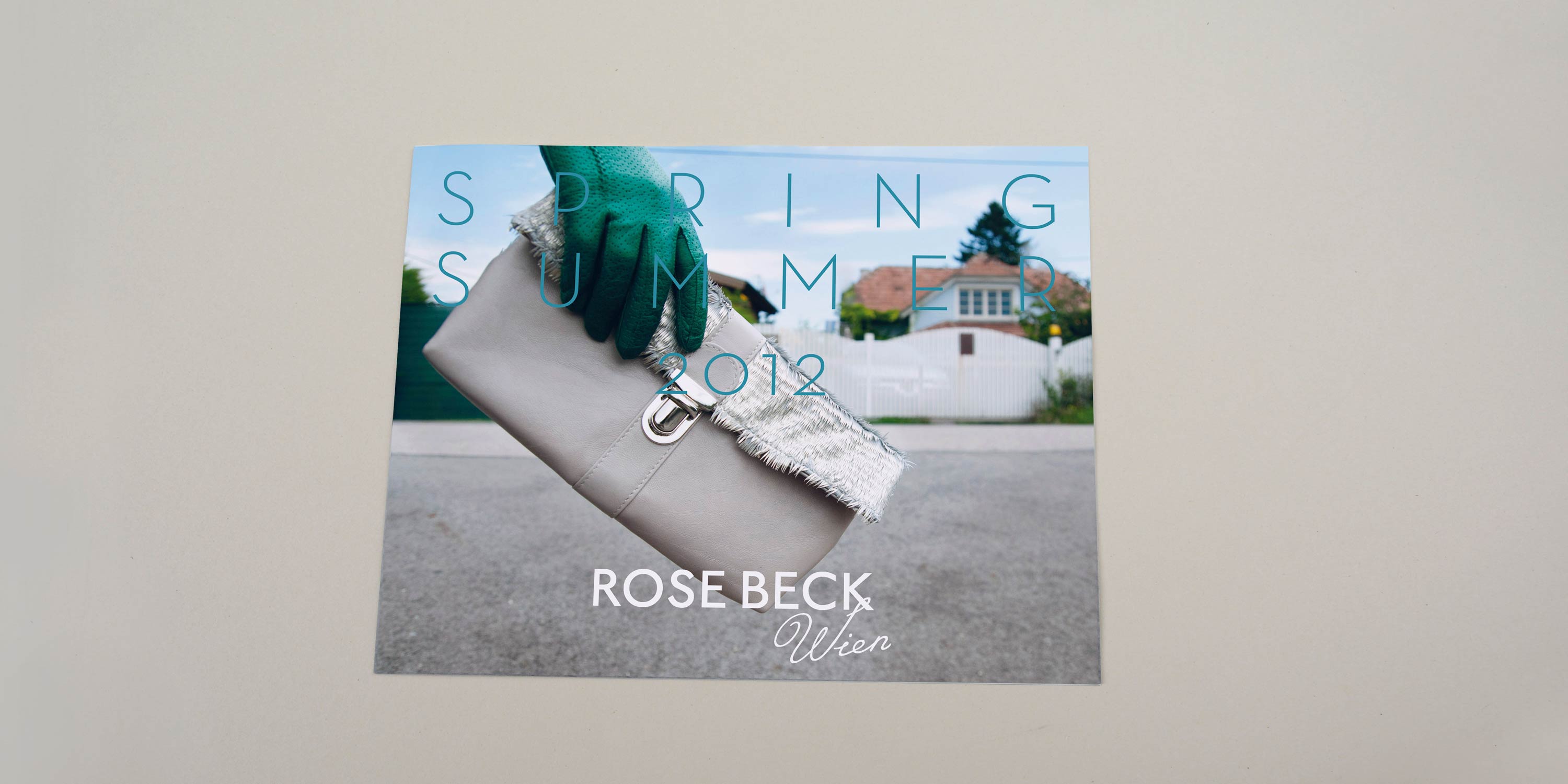 Cover catalogue. Close-up of hand holding handbag. Fence and houses in the background. Large title font  with lots of spacing overlayed. Logo at bottom overlayed.