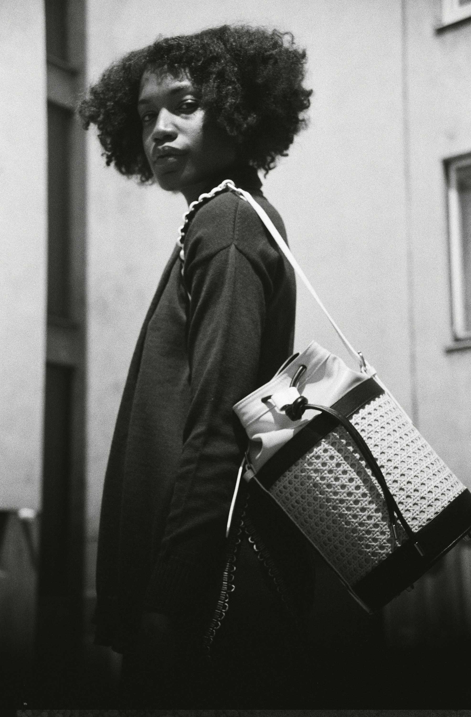 B/W photo. Woman in front of building. Carries handbag around her shoulder.