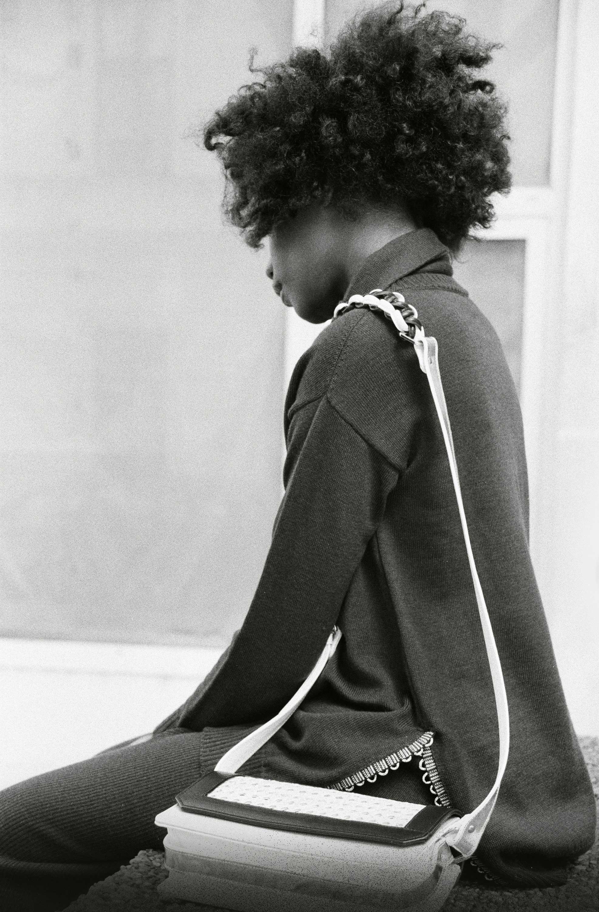 B/W photo. Woman sitting on concrete wall. Carries handbag around her shoulder. Building in the background.