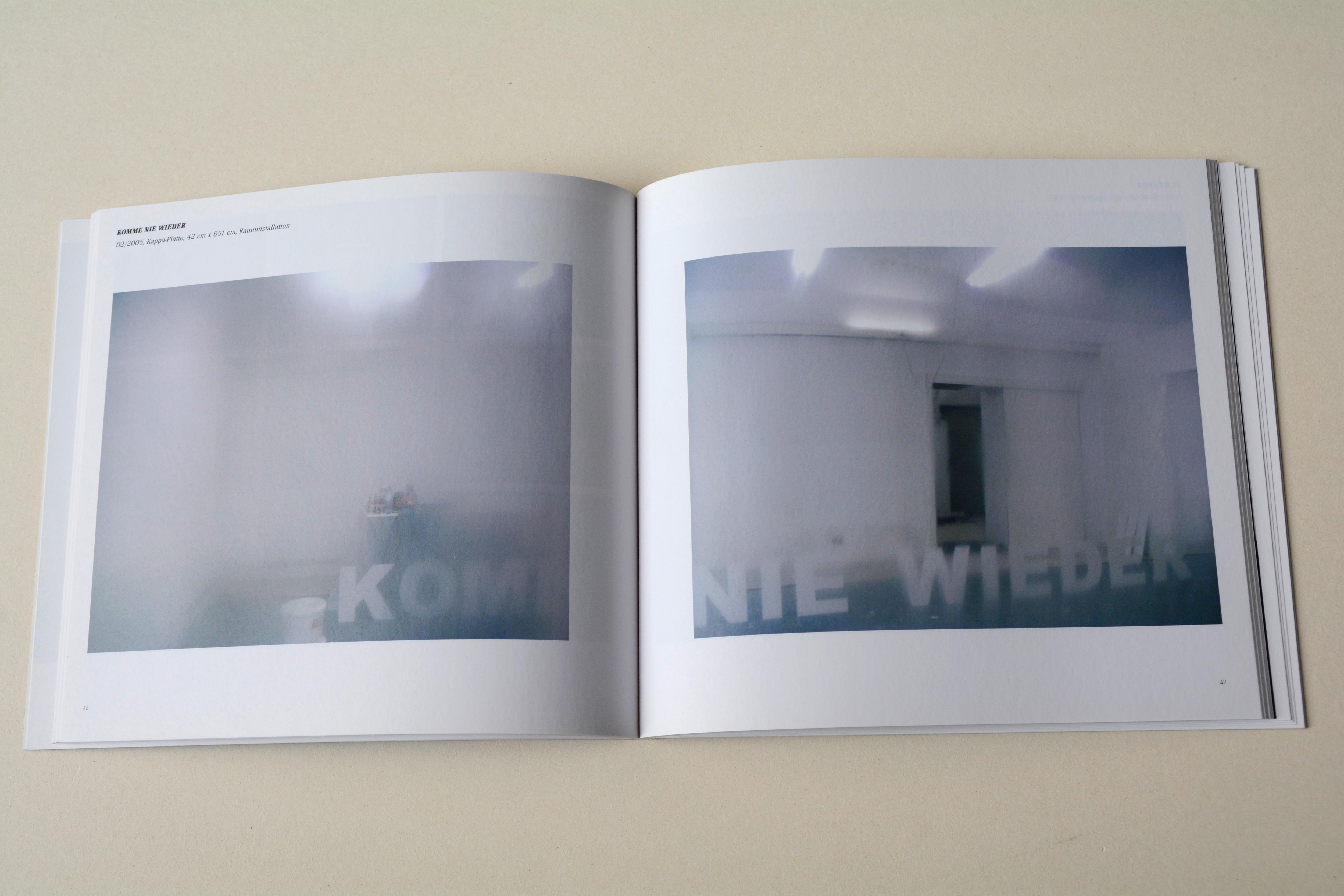 Double page. One blurry photo on each page. Line of small Text top left page. Left: White room with blue floor. Large letters on the floor forming part of a sentence. Right: White room with blue floor. Door in the back. Large letters on the floor forming part of a sentence.