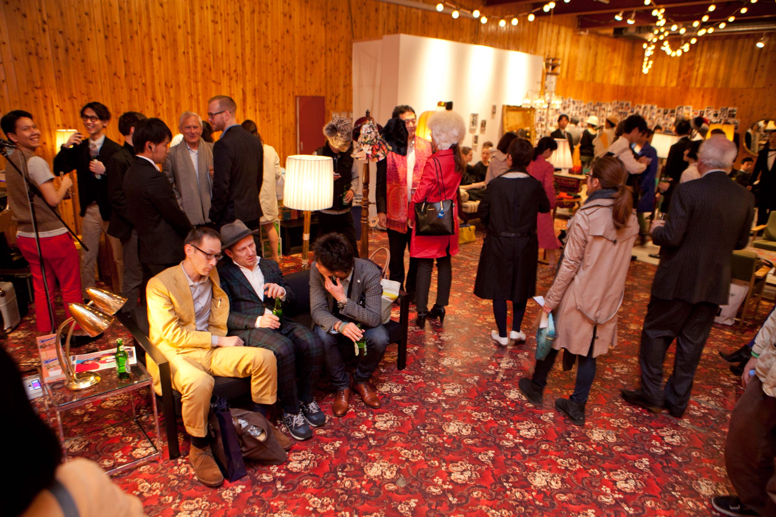 Crowded room. 3 persons sitting on couch. Others are standing. Walls covered with wooden panelling. Floor covered with ornamental pattern.